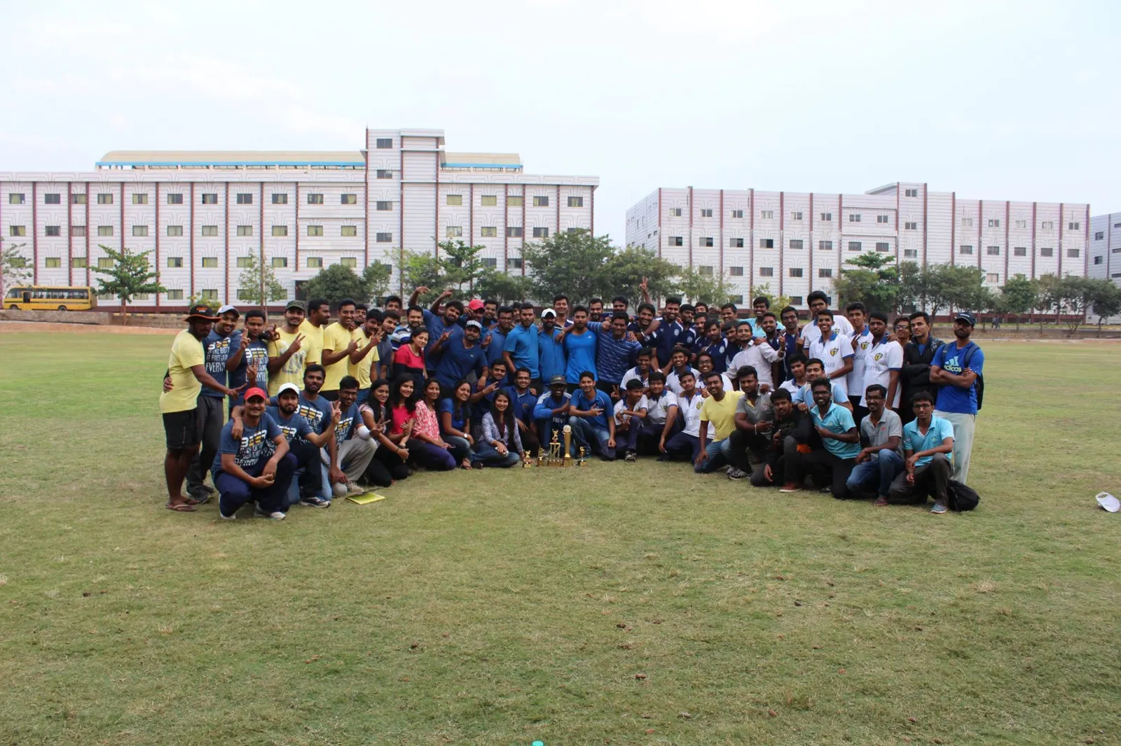 A large group photographed together on a sports ground, with trophies on the grass in front.