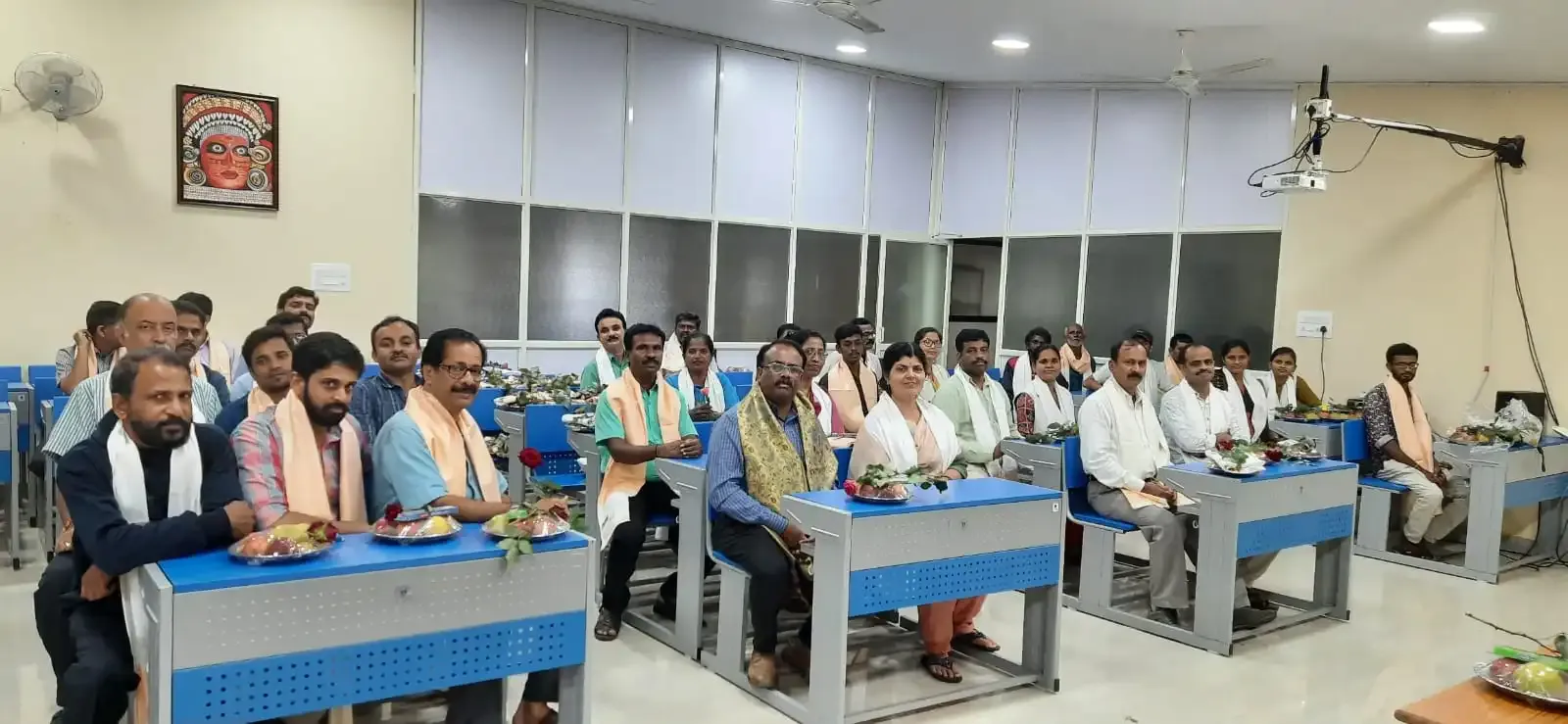 Teachers and guests seated at desks, wearing shawls presented during a felicitation.
